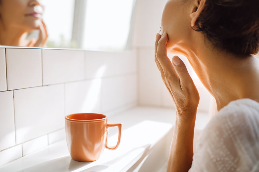 Mujer examinando su cuello frente al espejo del baño con taza de té, momento de autoconciencia corporal durante la menopausia